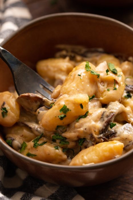 A bowl of creamy gnocchi topped with chopped herbs and mushrooms, with a fork holding a piece of gnocchi.