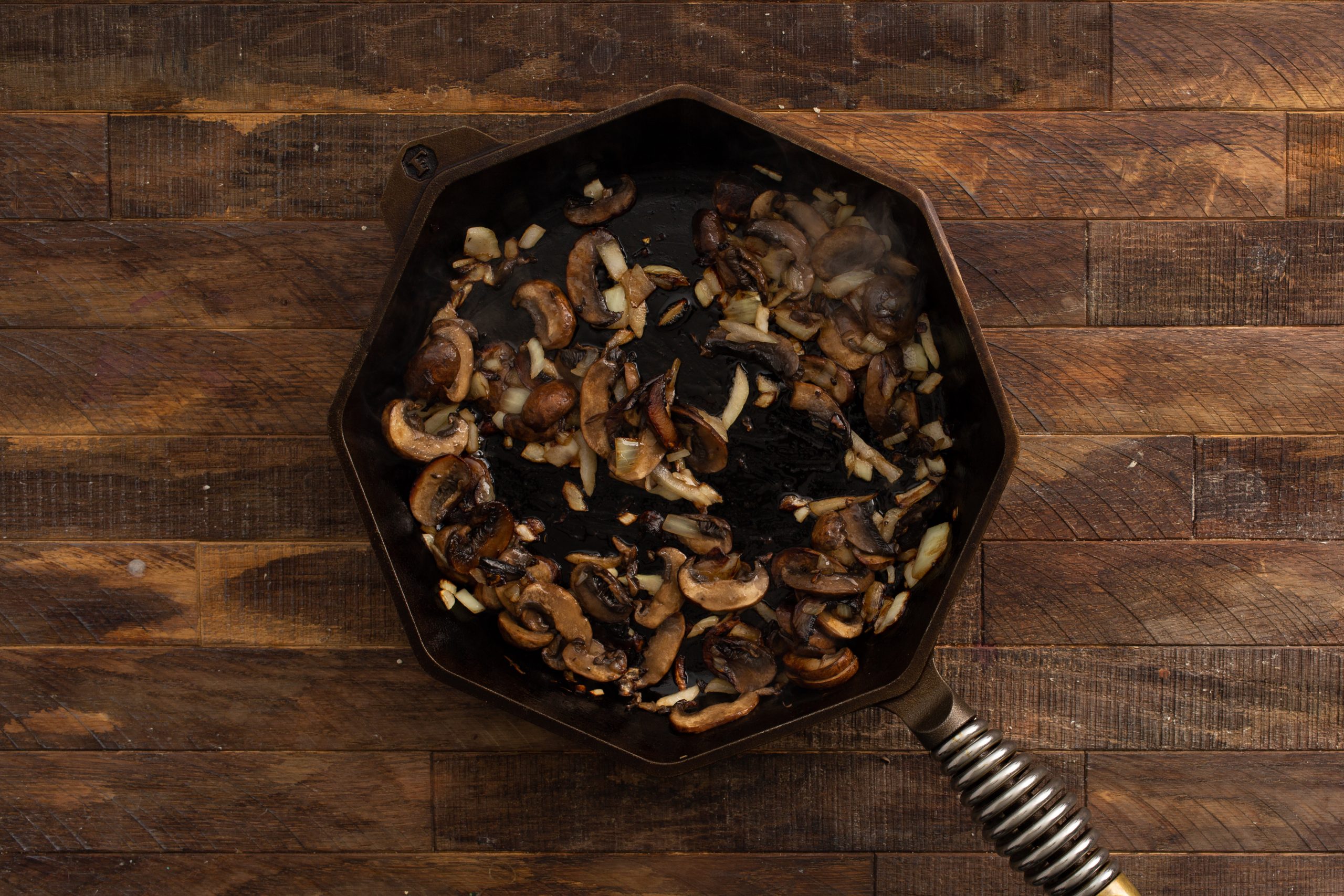 A hexagonal cast iron skillet placed on a wooden surface, containing sautéed mushrooms and onions.