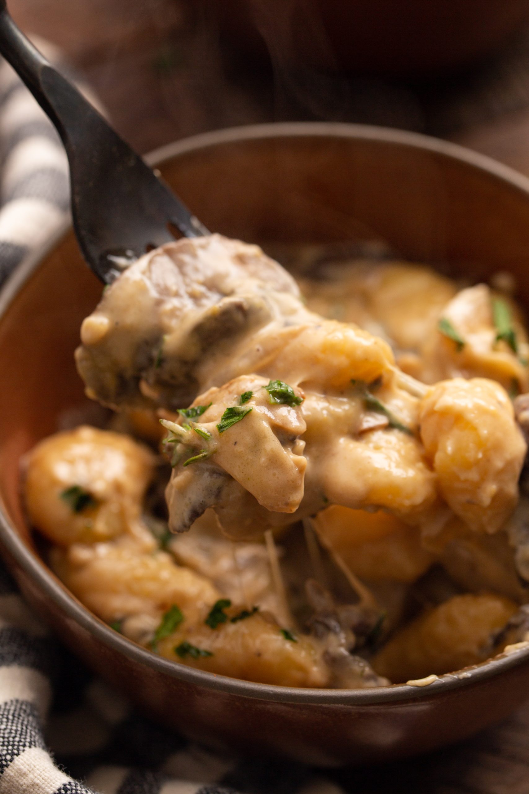 A close-up of gnocchi in a creamy sauce on a fork, garnished with herbs, with more gnocchi in a bowl underneath.