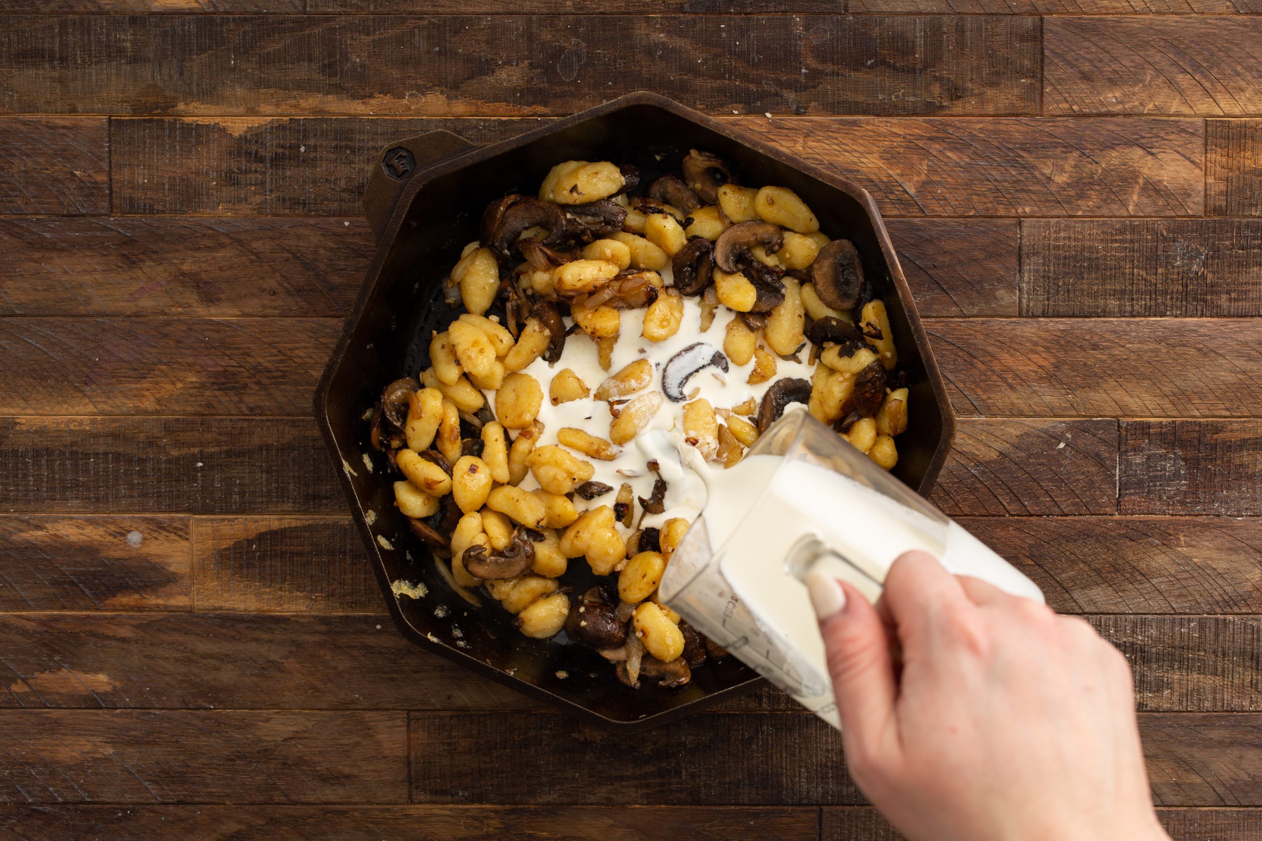 A hand pours cream from a glass container into a pan containing cooked gnocchi and mushrooms.