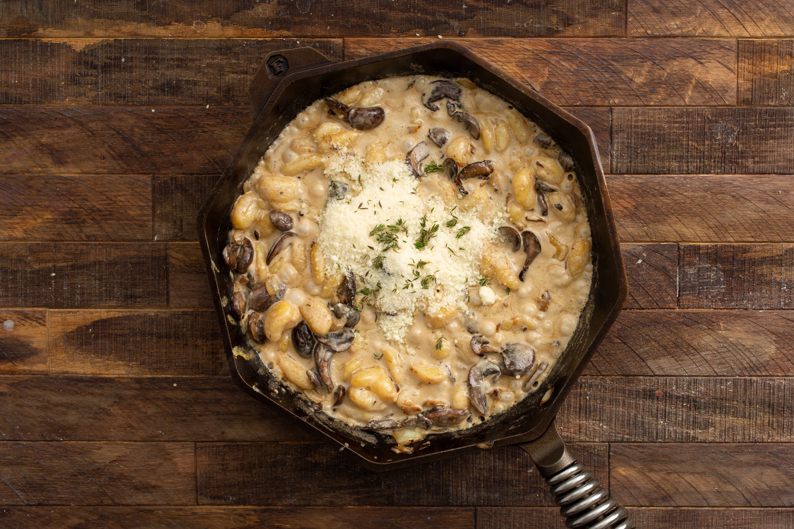 A skillet filled with creamy mushroom gnocchi topped with grated cheese and herbs, set on a wooden surface.