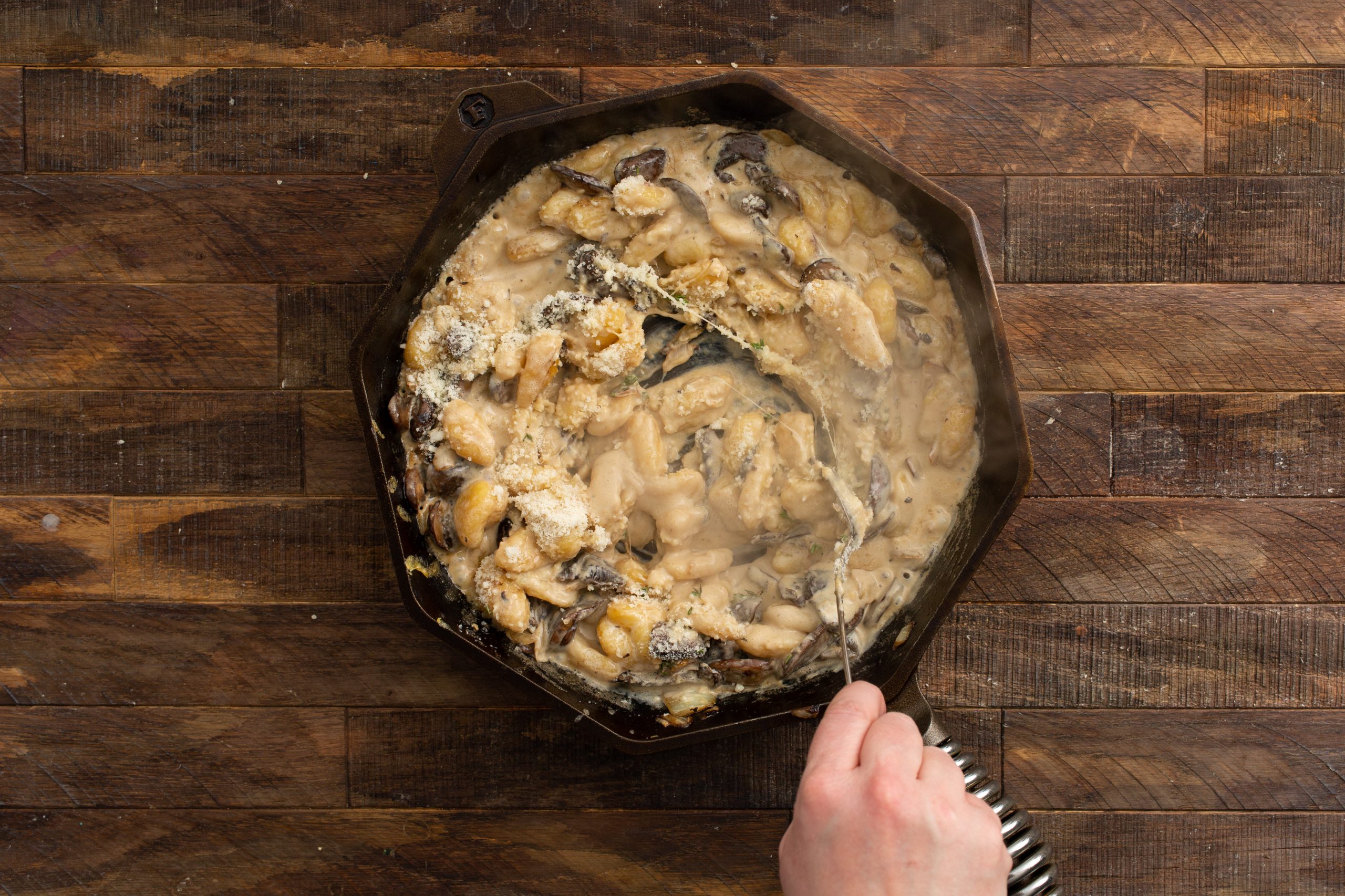 A hand stirring creamy mushroom dish in a hexagon-shaped pan on a wooden surface.