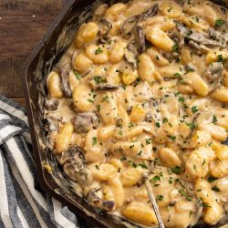 A cast iron skillet with creamy mushroom gnocchi garnished with chopped parsley, placed on a wooden table with a black and white striped cloth.