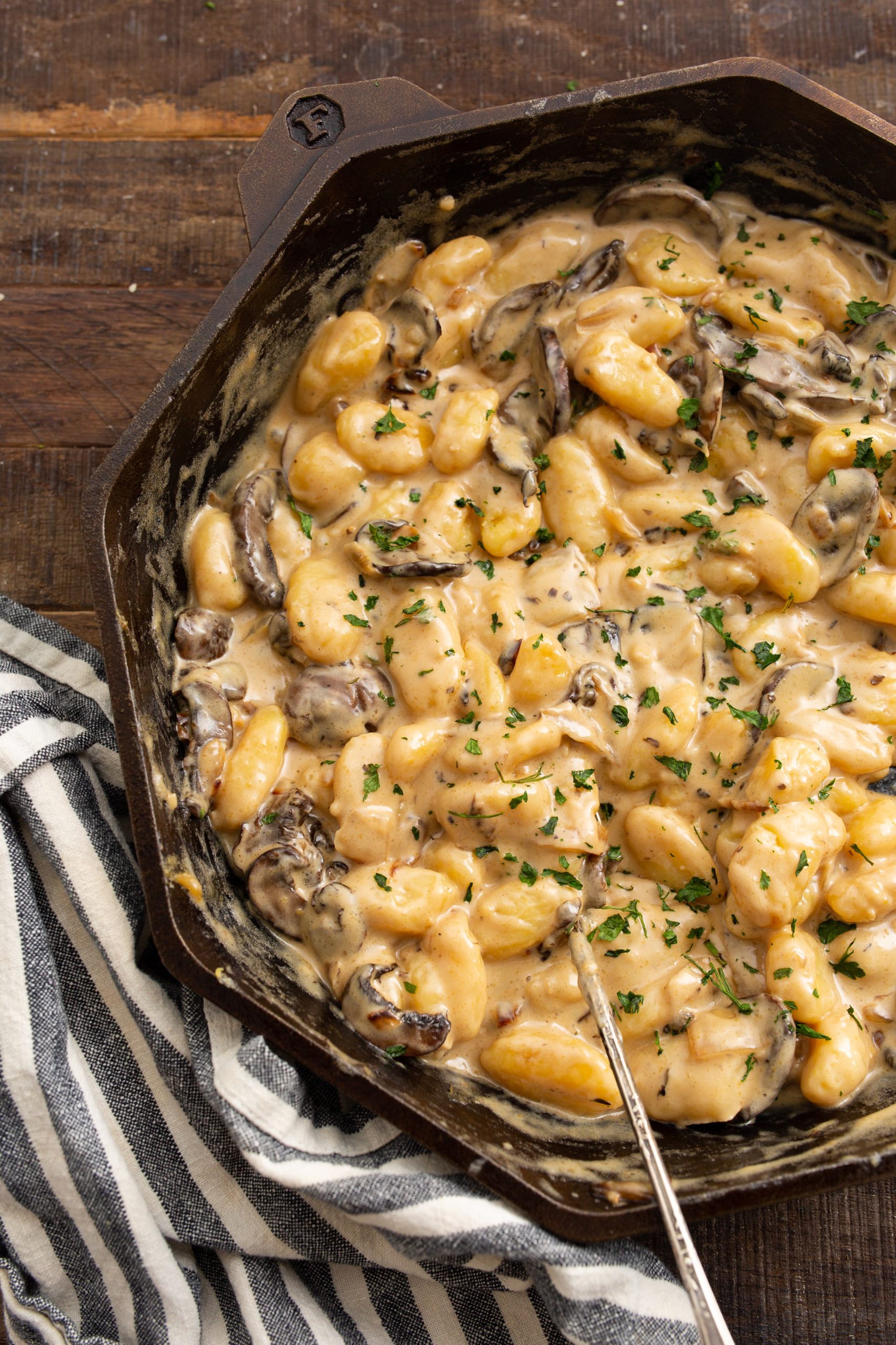 A cast iron skillet with creamy mushroom gnocchi garnished with chopped parsley, placed on a wooden table with a black and white striped cloth.