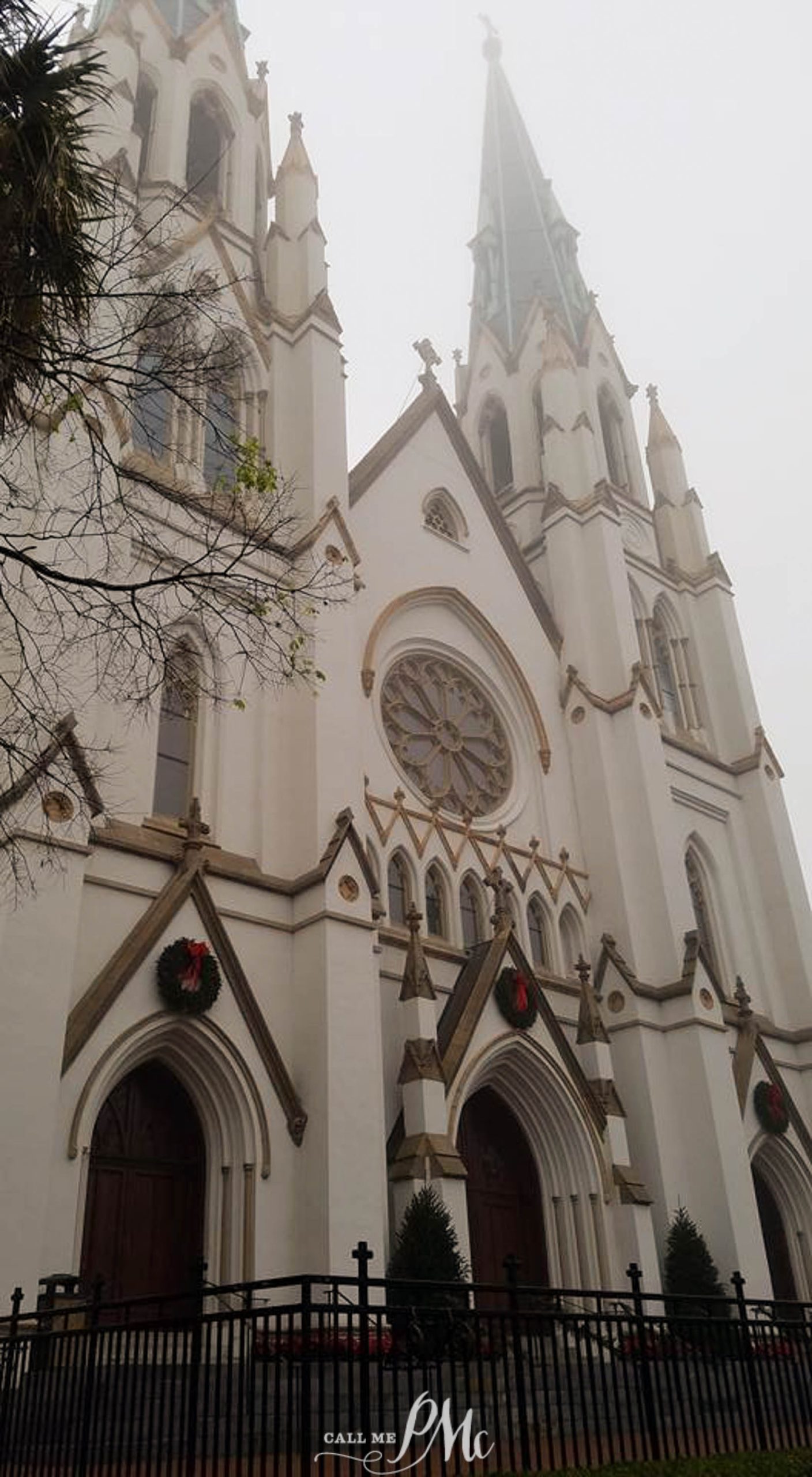 Image of a gothic-style cathedral with pointed arches, a large rose window, and twin towers rising into a foggy sky. The building is adorned with red and green Christmas wreaths.