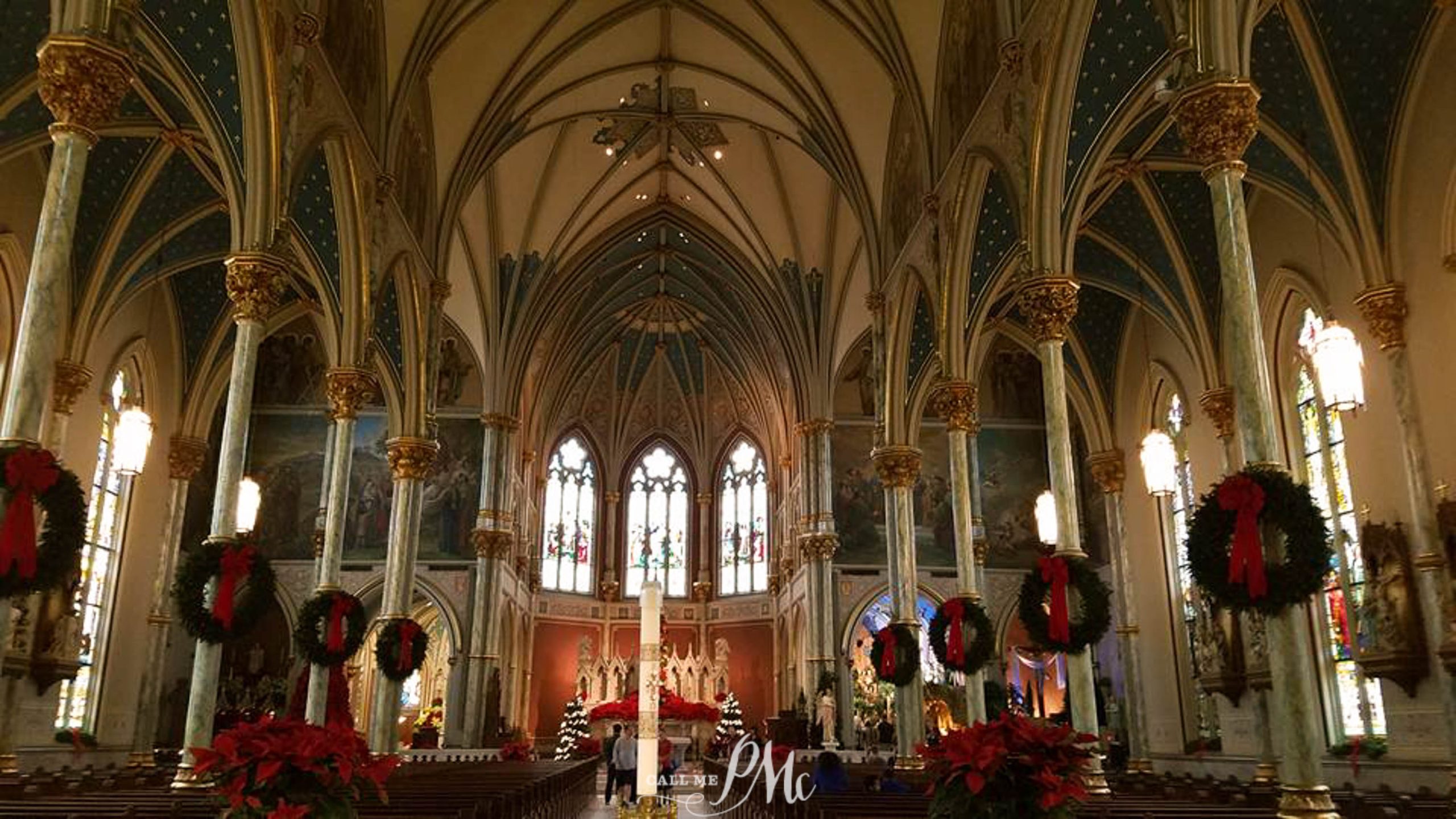 The image shows the interior of a decorated cathedral with high vaulted ceilings, stained glass windows, and Christmas wreaths hanging from the columns.