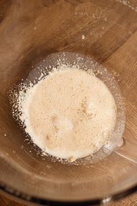 A close-up of a glass bowl containing a frothy mixture of yeast and water with bubbles forming on the surface, set on a wooden countertop.