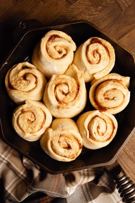 A cast iron skillet filled with seven uncooked cinnamon rolls, arranged in a circular pattern, resting on a checkered cloth.