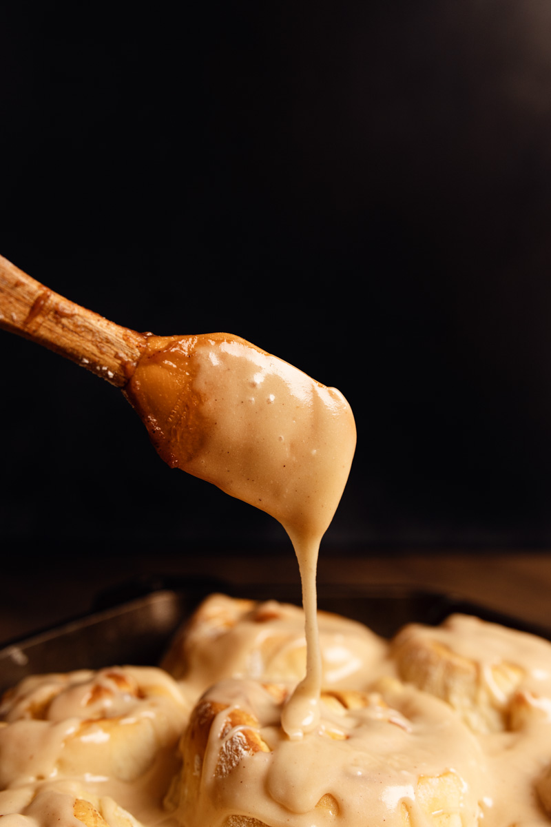 A wooden spoon drizzles creamy icing over freshly baked cinnamon rolls in a dark background.