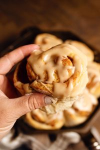 A hand holding a frosted cinnamon roll above a tray of more cinnamon rolls. The rolls have a creamy glaze and are in focus against a blurred background.