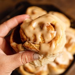 A hand holding a frosted cinnamon roll above a tray of more cinnamon rolls. The rolls have a creamy glaze and are in focus against a blurred background.