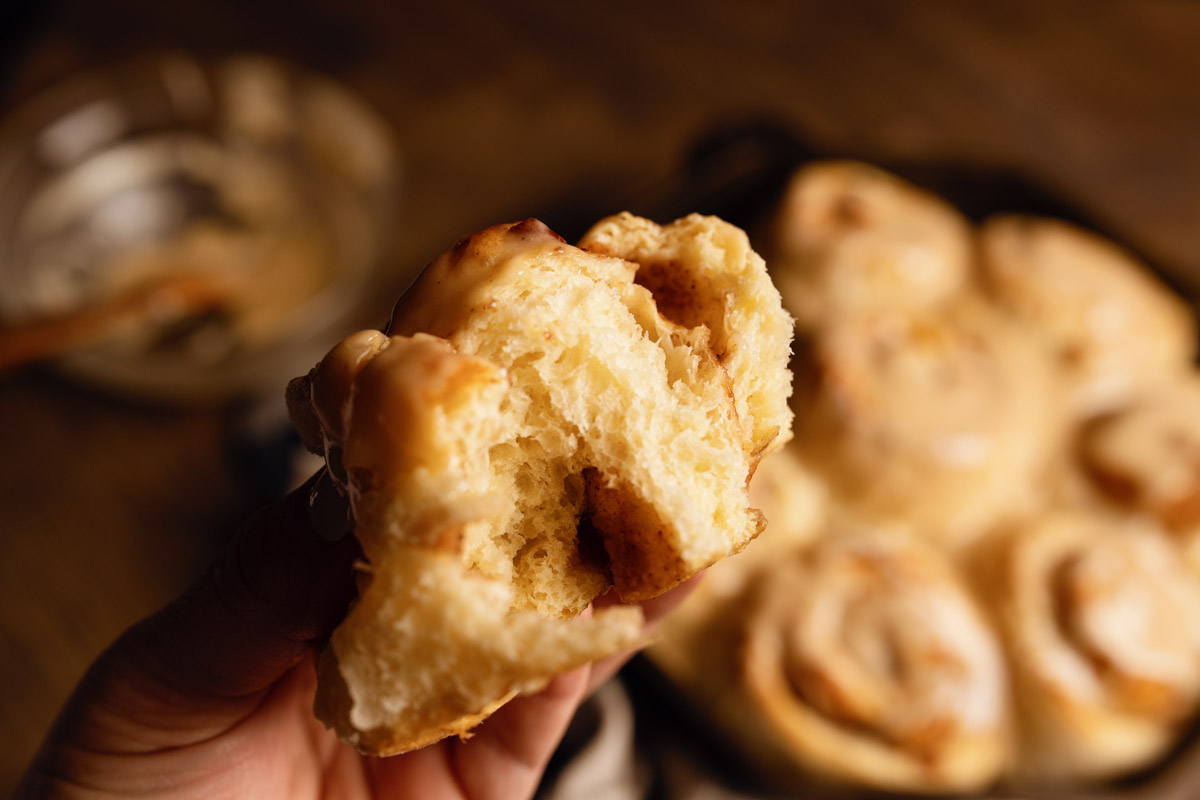 A close-up of a hand holding a partially eaten cinnamon roll with more rolls in the background.