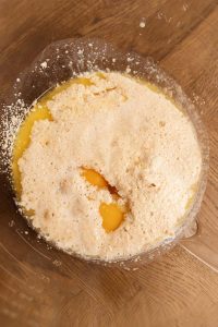 A clear bowl filled with frothy milk, melted butter, and two egg yolks on a wooden surface.