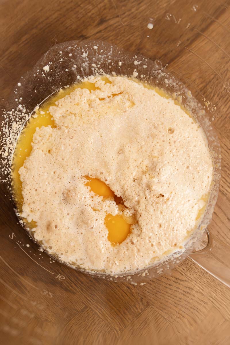 A clear bowl filled with frothy milk, melted butter, and two egg yolks on a wooden surface.