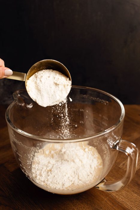 A hand holding a measuring cup pours flour into a glass mixing bowl already containing some flour, set on a wooden surface against a dark background.