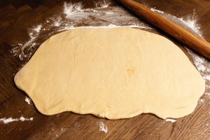 A rolled-out piece of dough on a floured surface with a wooden rolling pin nearby.