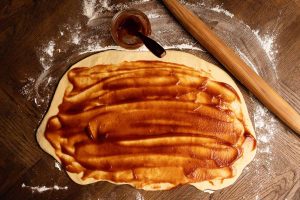 Rolled-out dough on a floured surface spread with brown sauce, next to a rolling pin and a small bowl containing more sauce.