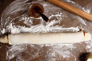 A rolled-out dough with filling sits on a floured wooden surface. A rolling pin and a small bowl with a spoon containing a brown substance are placed nearby.