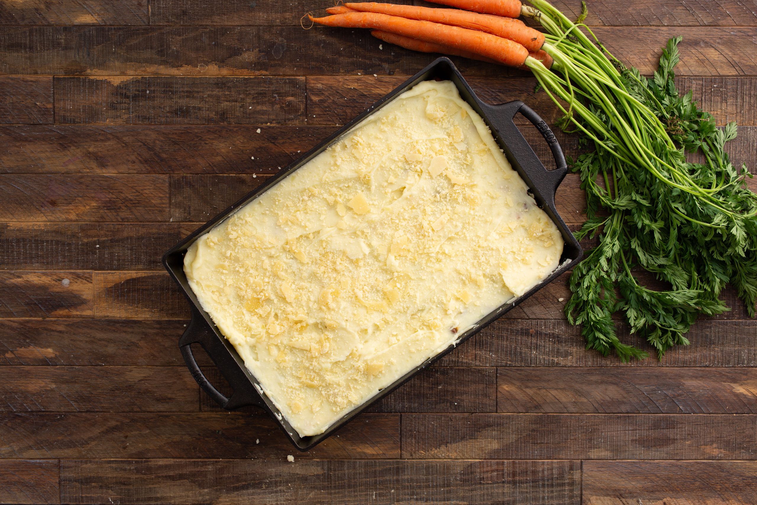 A rectangular casserole dish filled with a baked dish topped with grated cheese or crumbs, next to a bunch of fresh carrots with leafy greens on a wooden surface.