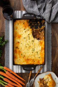 A rectangular dish containing a partially-served shepherd's pie made from an authentic cottage pie recipe. Surrounding the dish are a glass of wine, carrots, a plate with a serving, utensils, and a gray cloth on a wooden surface.