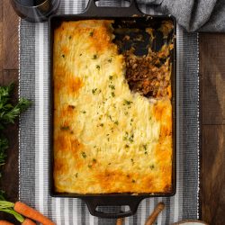 A rectangular dish containing a partially-served shepherd's pie made from an authentic cottage pie recipe. Surrounding the dish are a glass of wine, carrots, a plate with a serving, utensils, and a gray cloth on a wooden surface.