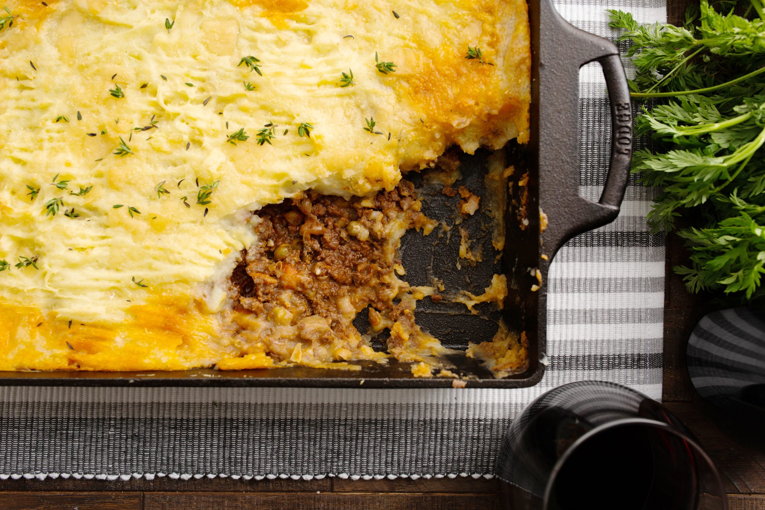 A partially eaten Authentic Cottage Pie Recipe in a baking dish on a checkered tablecloth, with a glass of red wine and sprigs of parsley beside it.