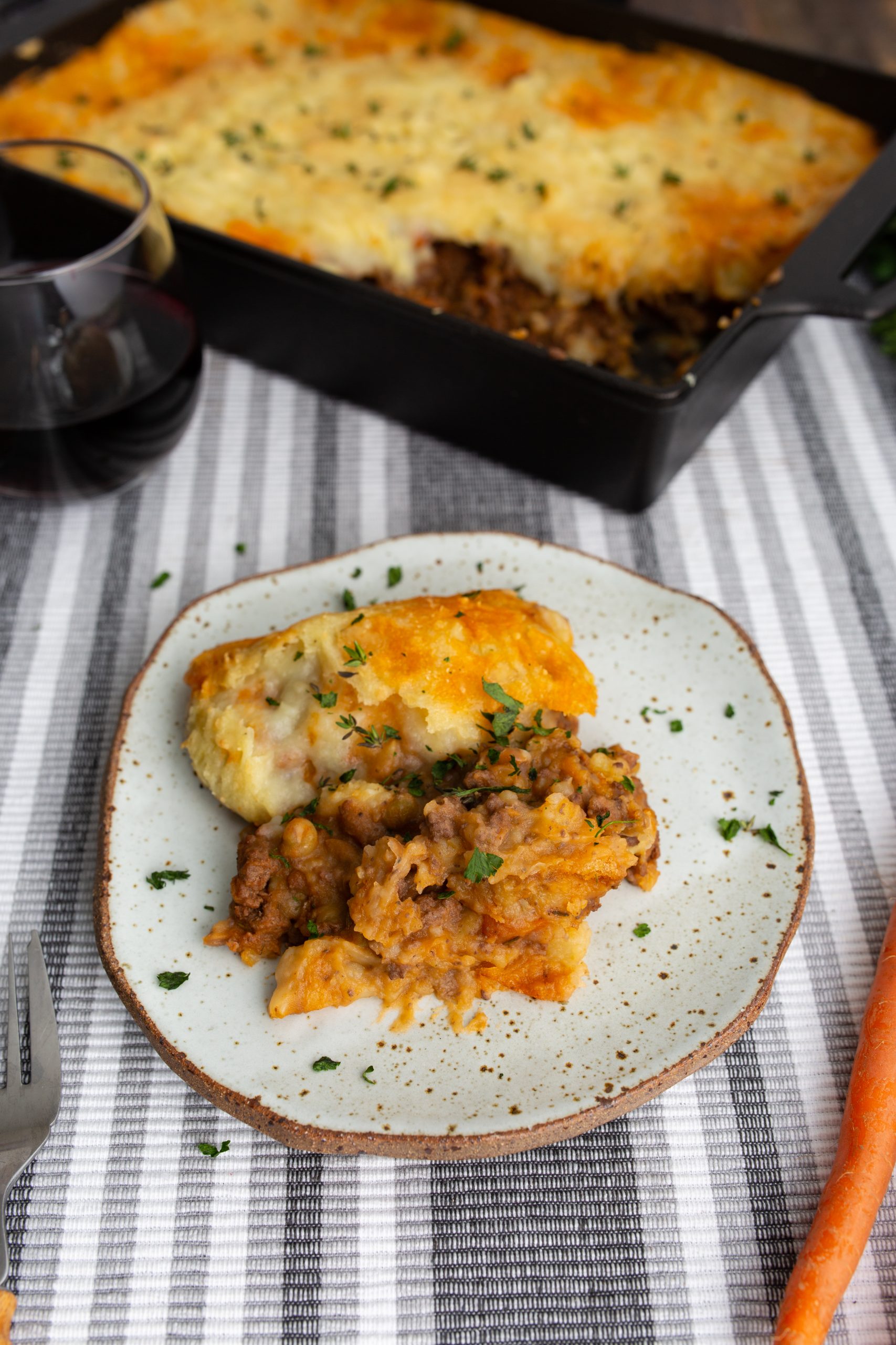 A plate of shepherd's pie garnished with herbs sits on a striped placemat, with a baking dish of shepherd's pie and a glass of red wine in the background.