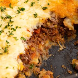 A close-up of a partially eaten shepherd's pie in a baking dish, showing layers of mashed potatoes, ground beef, and vegetables, topped with melted cheese and garnished with chopped parsley.