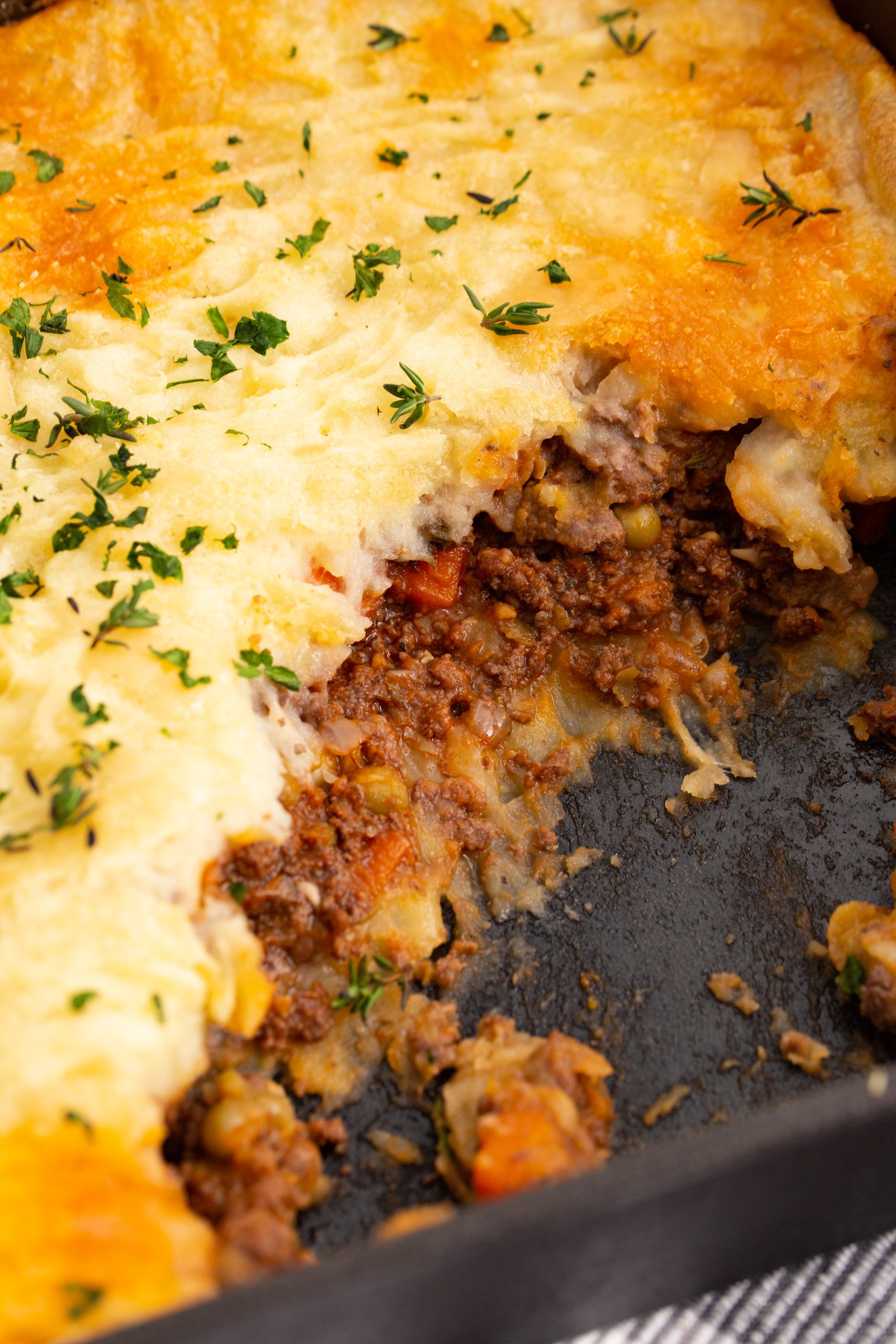 A plate of cottage pie garnished with herbs sits on a striped placemat, with a baking dish of shepherd's pie and a glass of red wine in the background.