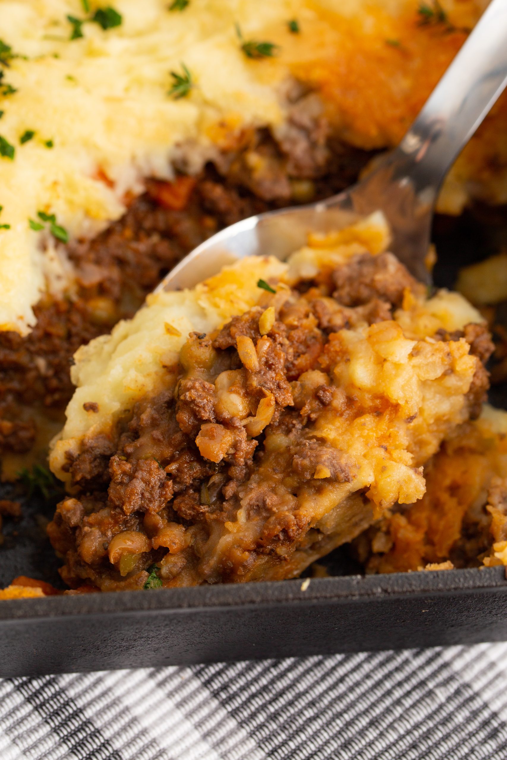 A spoon scooping a portion of Authentic Cottage Pie Recipe from a dish. The pie has a mashed potato topping with ground meat and vegetable filling underneath.