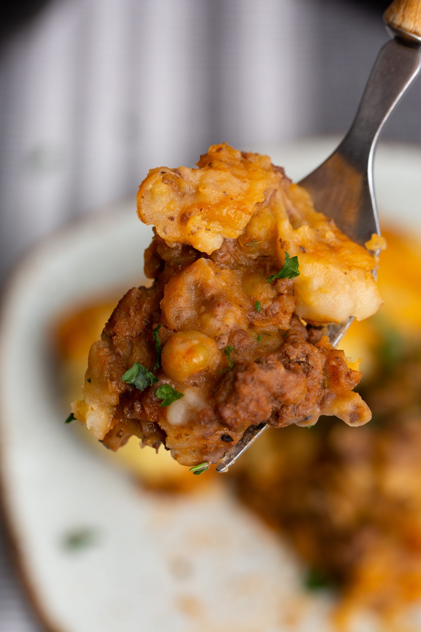 Close-up of a fork holding a bite of shepherd's pie with visible meat, vegetables, and mashed potatoes, garnished with parsley.