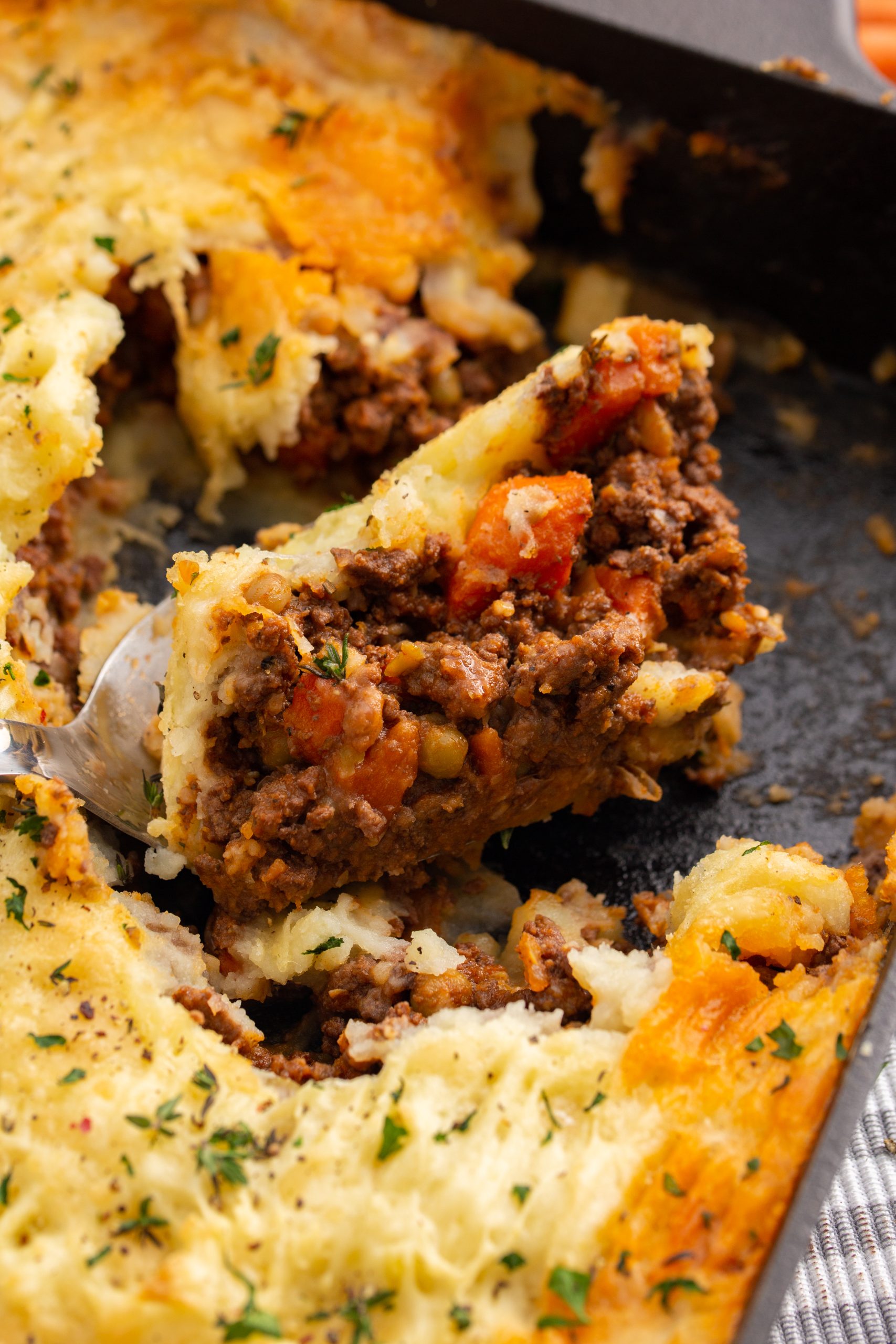 A close-up of a slice of Authentic Cottage Pie Recipe being lifted out of a dish, showing layers of mashed potatoes, ground meat, and vegetables. The dish is garnished with chopped herbs.