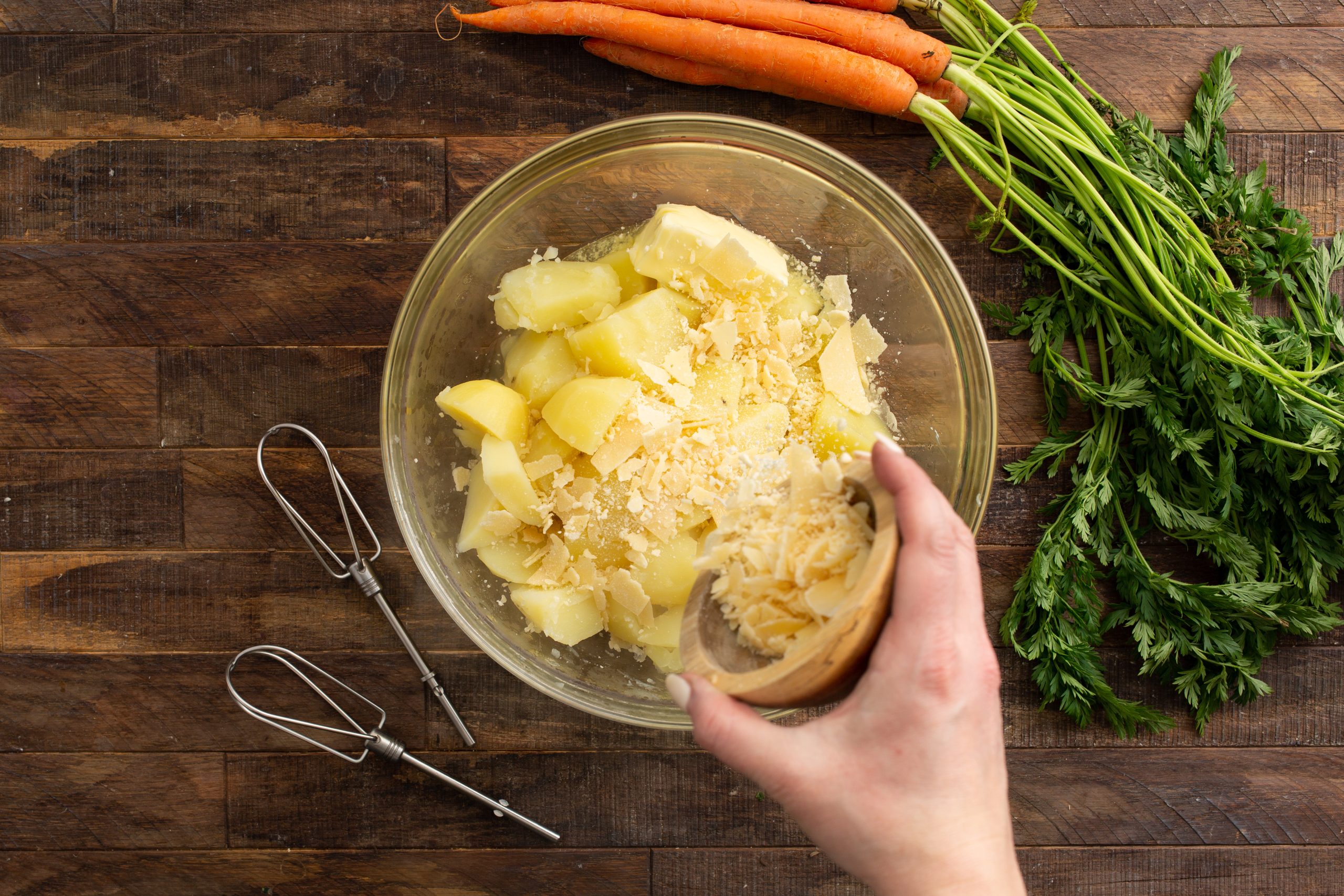 A hand is adding grated cheese to a bowl of cooked potatoes. Mixer attachments and fresh carrots with greens are on a wooden surface.