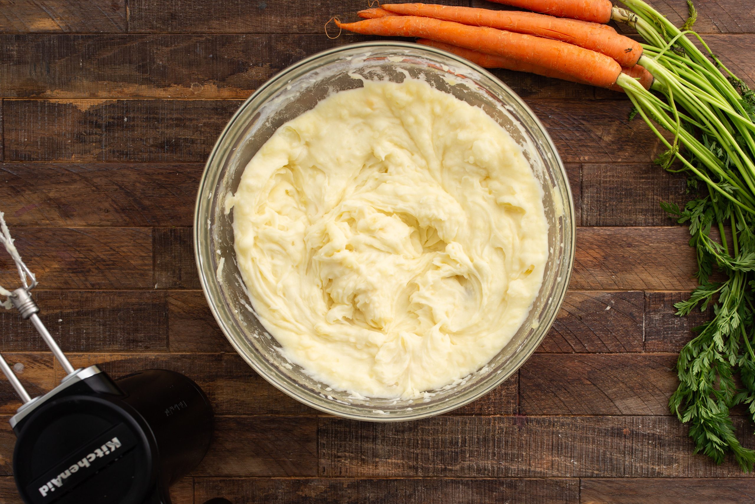 A bowl of mashed potatoes on a wooden surface with a hand mixer and several fresh carrots with greens nearby.