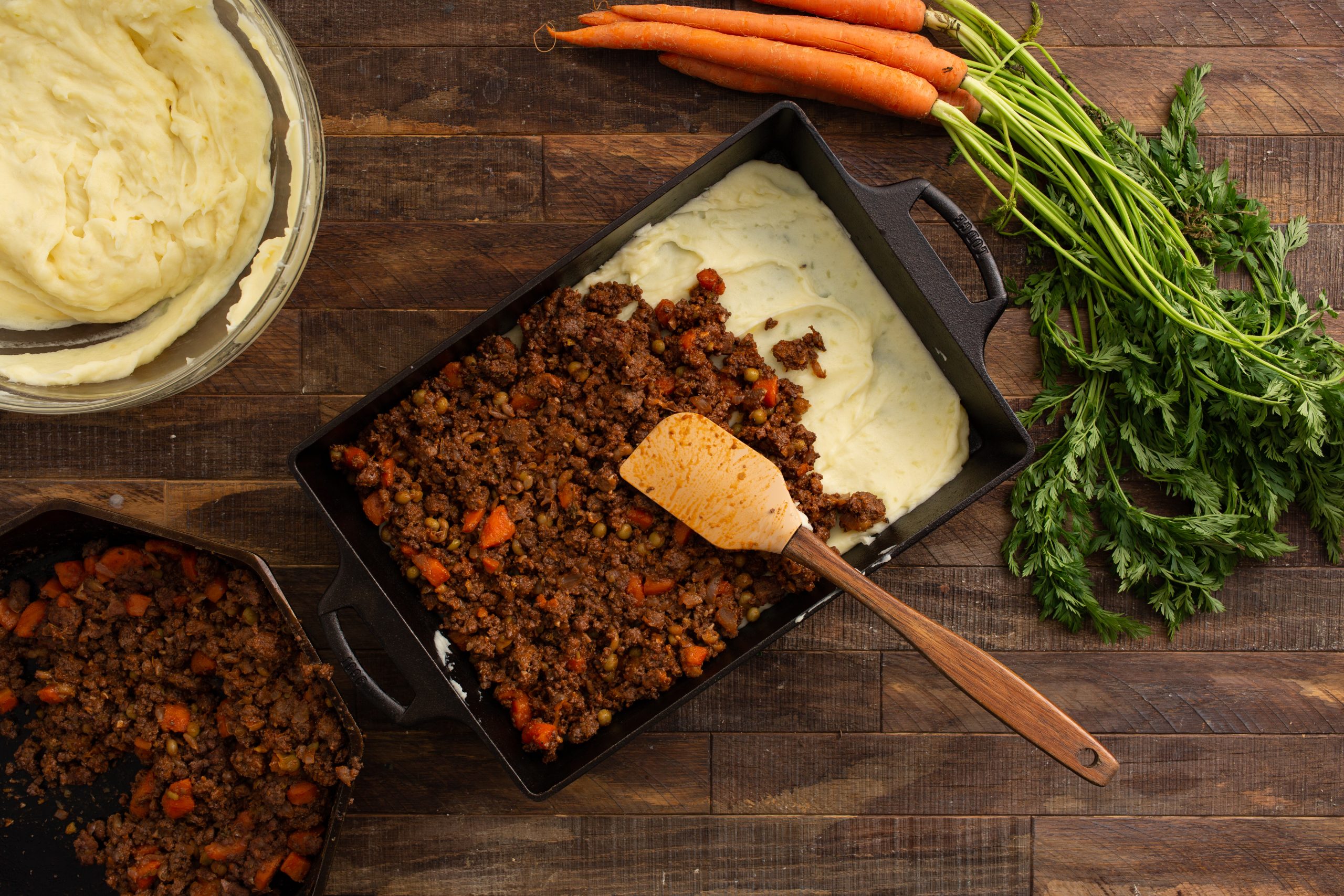 A casserole being prepared with layers of ground meat and mashed potatoes in a black baking dish, surrounded by fresh carrots and greens on a wooden surface.