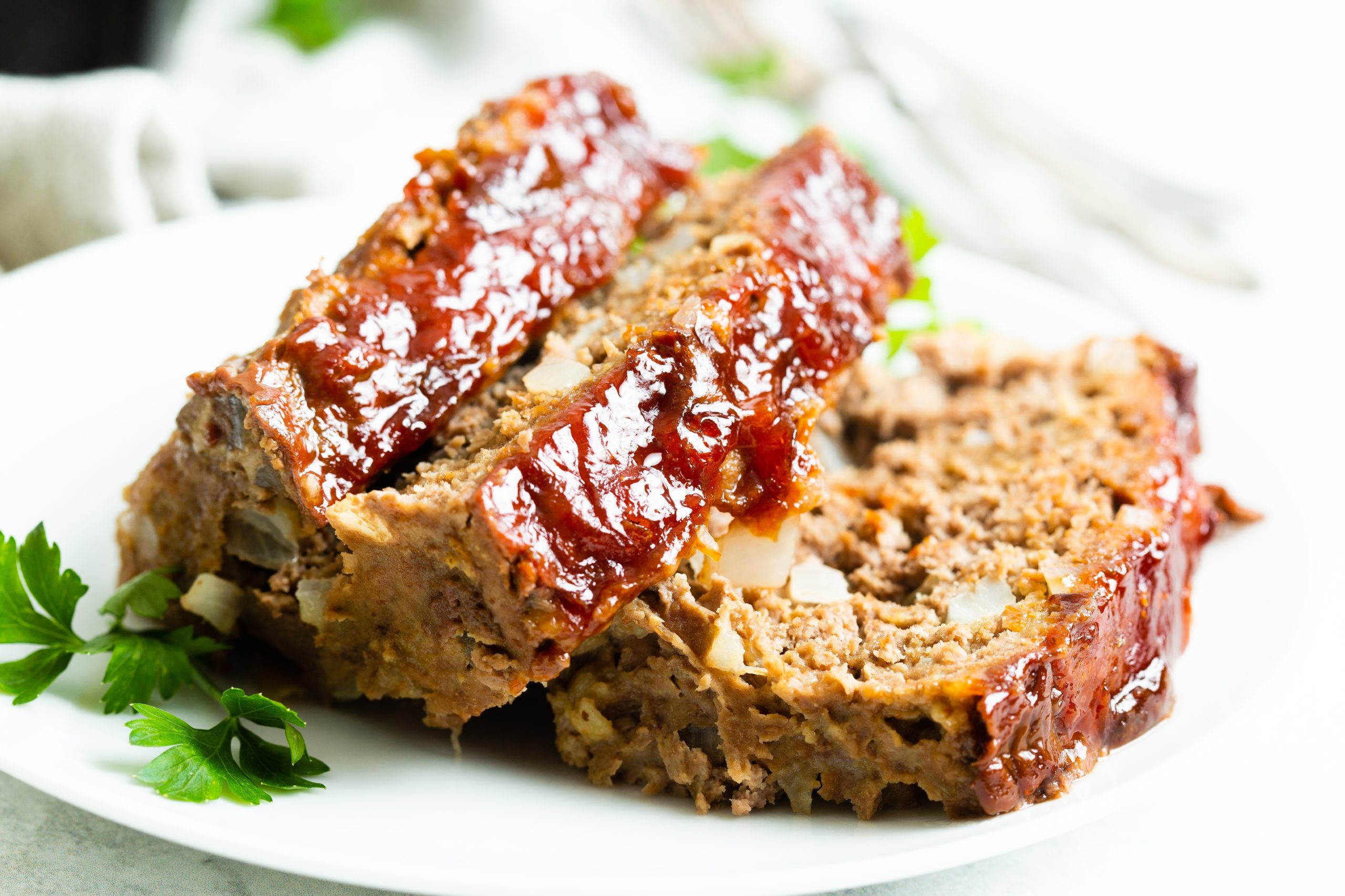 Close-up of two slices of glazed meatloaf on a white plate, garnished with fresh parsley. A fork and knife are blurred in the background.