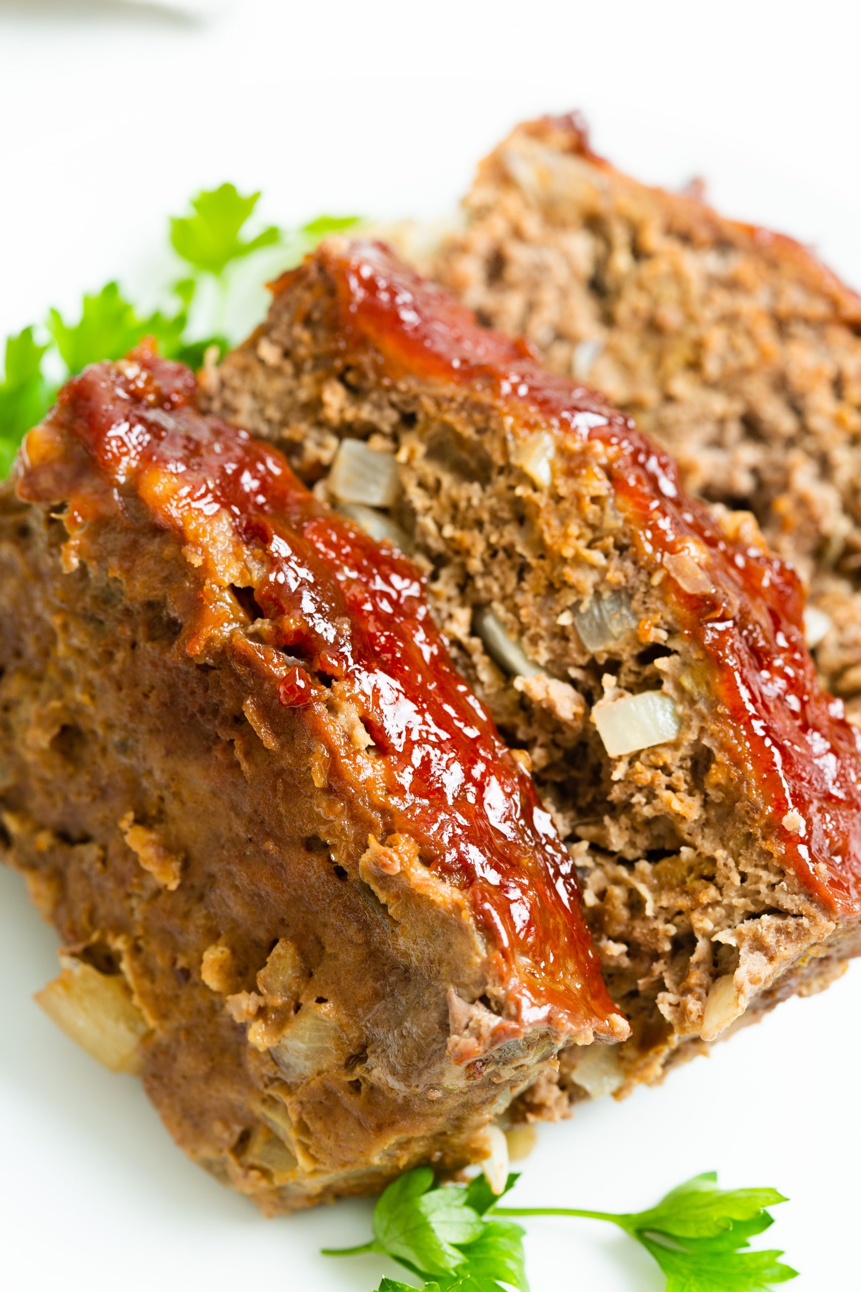 Close-up of two slices of glazed meatloaf on a white plate, garnished with fresh parsley. A fork and knife are blurred in the background.