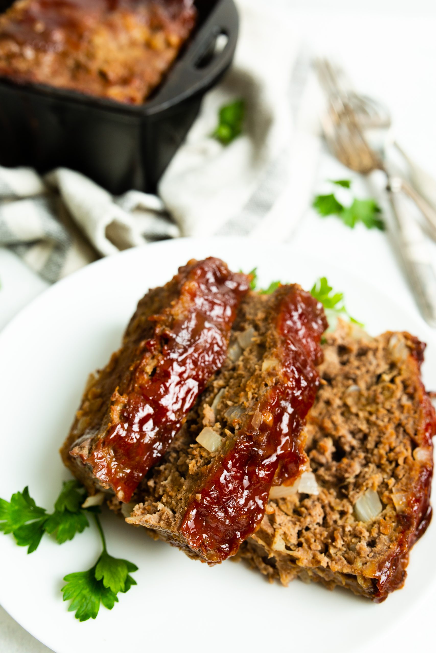 A close-up of two slices of seasoned Cast Iron Loaf Pan Meatloaf with a shiny glaze, served on a white plate garnished with parsley. A cast iron dish with more meatloaf is in the background.