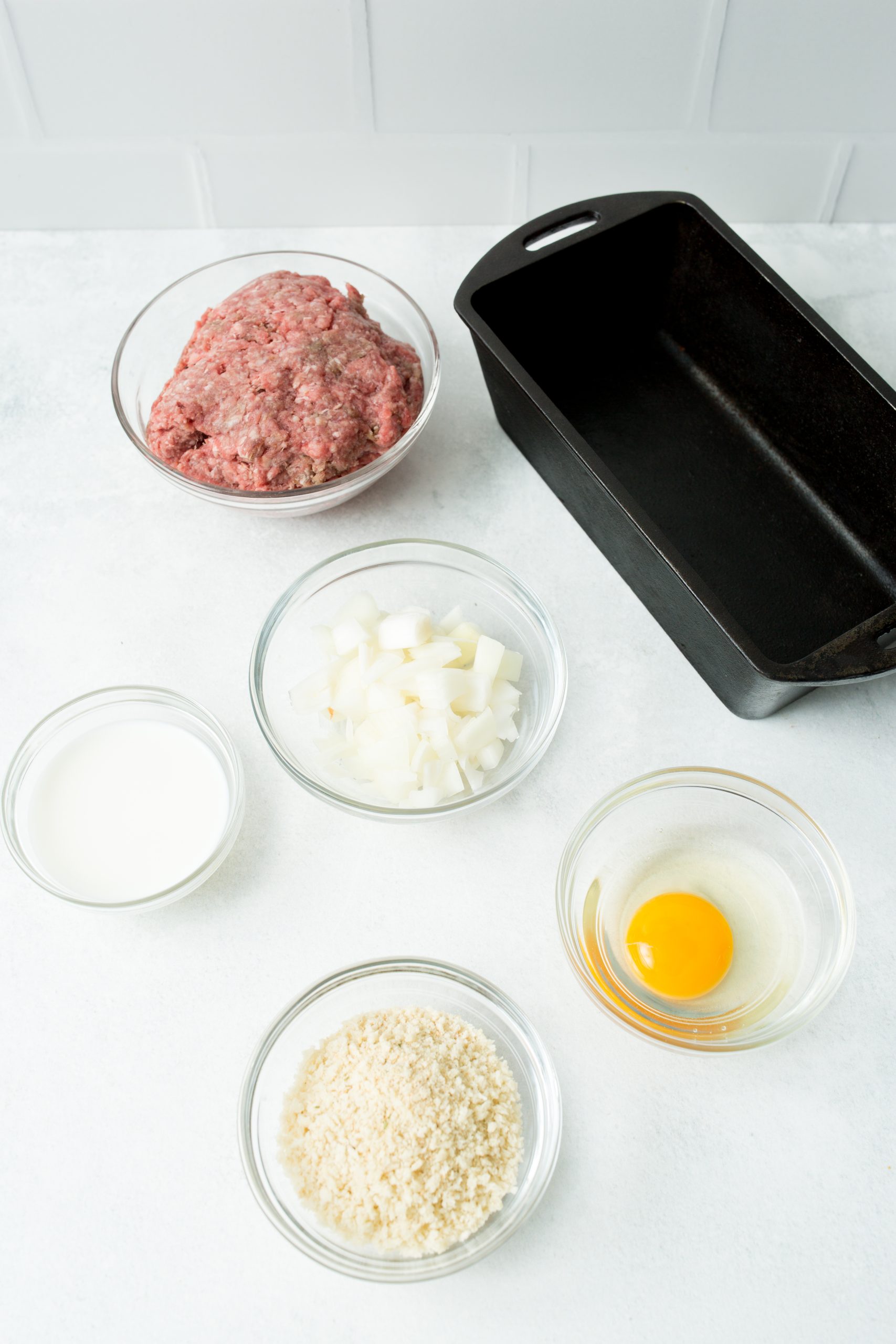 Ingredients for meatloaf arranged on a countertop: ground meat, chopped onions, an egg, breadcrumbs, and milk. A rectangular empty baking dish is positioned beside the ingredients.