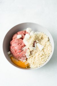 A bowl containing raw ground meat, chunks of white onion, breadcrumbs, and a cracked raw egg on a white surface.