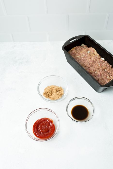 A Cast Iron Loaf Pan Meatloaf mixture in a black baking pan sits on a white countertop. Nearby are small bowls containing ketchup, brown sugar, and Worcestershire sauce.