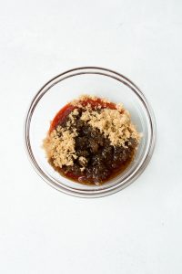 A glass bowl containing a mixture of brown sugar, soy sauce, and ketchup on a white surface.