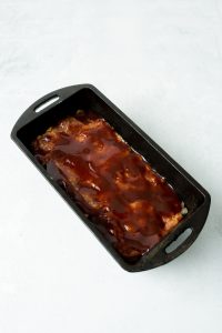 A rectangular black pan contains a meatloaf topped with a red sauce, set on a light gray surface.