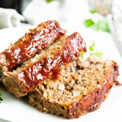 A plate with two slices of glazed meatloaf garnished with parsley, set on a white surface with a fork in the background.