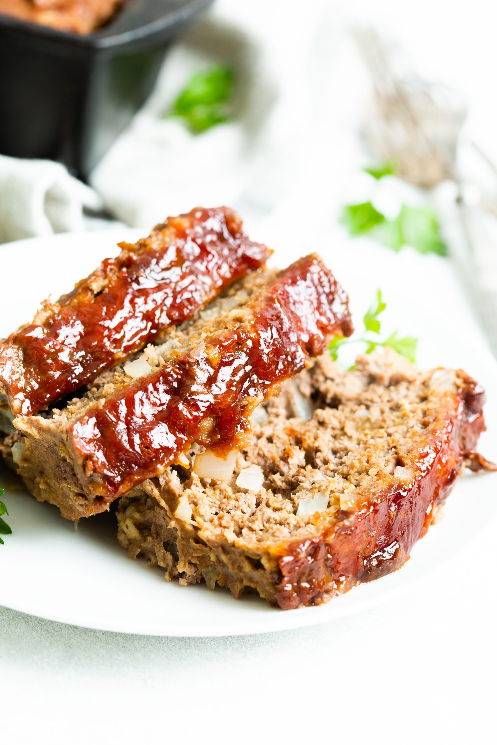 A close-up of two slices of seasoned Cast Iron Loaf Pan Meatloaf with a shiny glaze, served on a white plate garnished with parsley. A cast iron dish with more meatloaf is in the background.