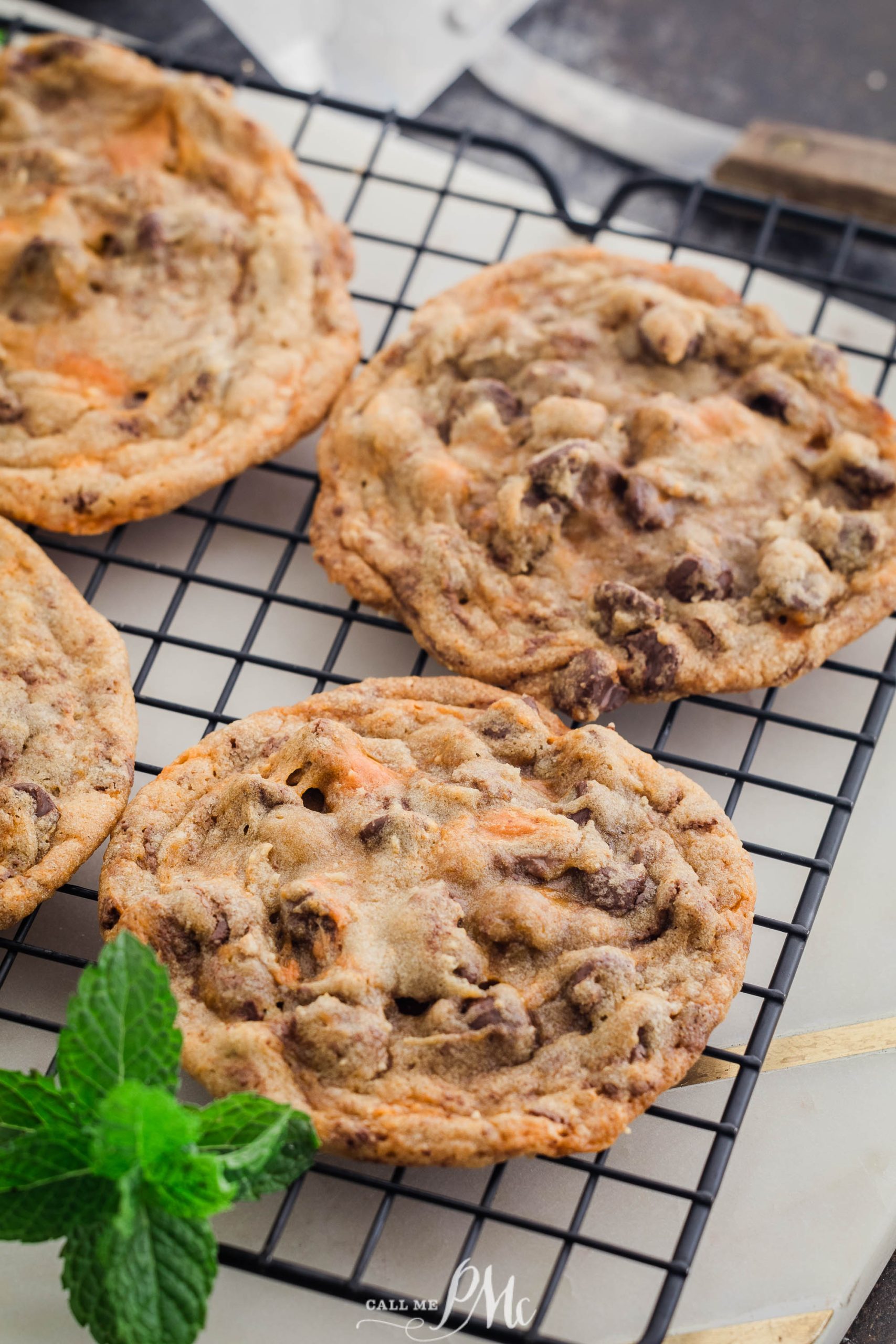 Four freshly baked chocolate chip cookies cool on a wire rack with a sprig of mint placed nearby.