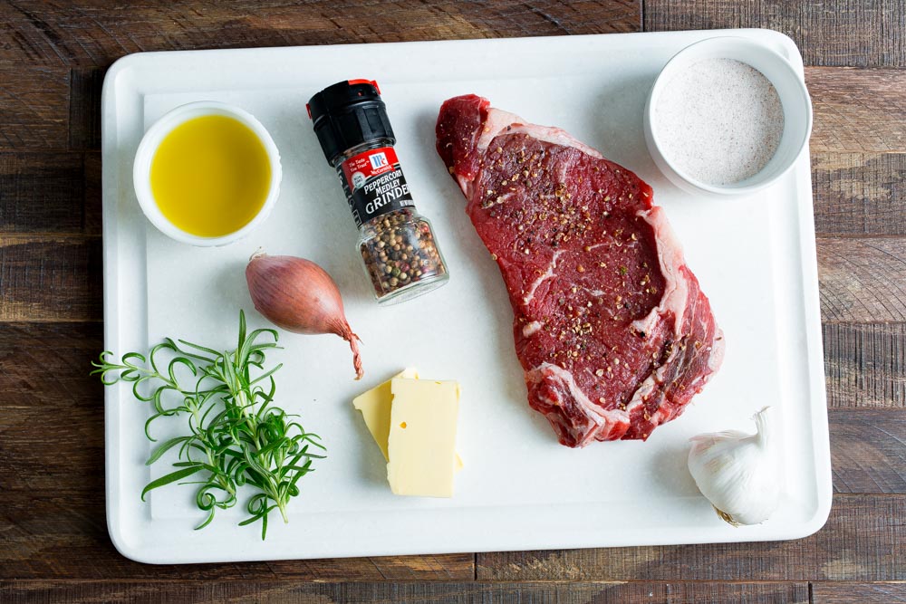 A white tray on a wooden surface holds a raw steak, a bowl of olive oil, salt, a pepper grinder, shallot, garlic clove, rosemary, and slices of butter.