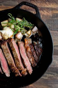 A cast-iron skillet with sliced steak, garnished with rosemary, roasted garlic, and charred shallots on a wooden surface.