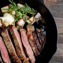 A cast-iron skillet with sliced steak, garnished with rosemary, roasted garlic, and charred shallots on a wooden surface.