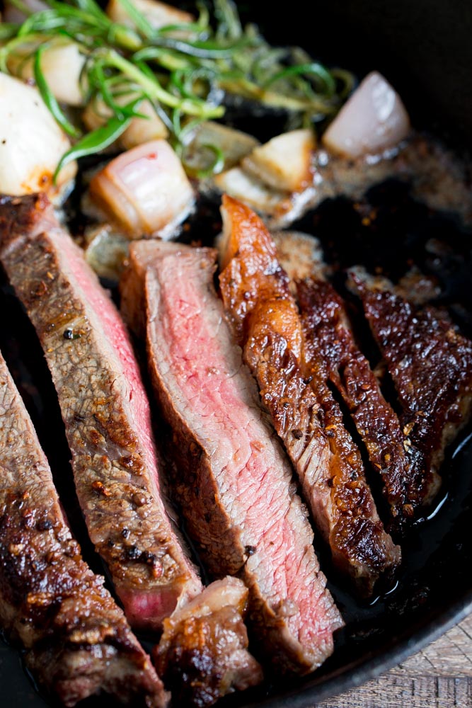 Close-up of sliced medium-rare steak with a seared crust, garnished with green herbs and cooked garlic in a cast-iron skillet.